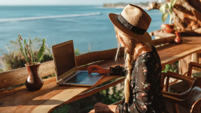 Mulher de chapéu e vestido floral trabalhando em laptop com vista para o mar azul em um balcão de madeira.