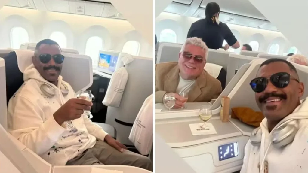 Milton Cunha e seu namorado sorrindo e brindando com taças de vinho no avião, em assentos de primeira classe.
