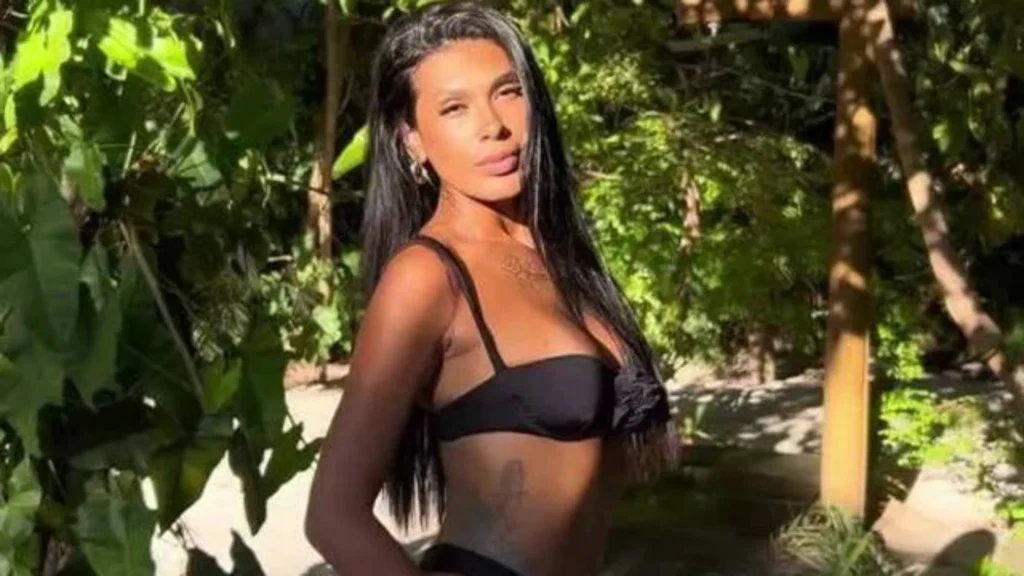 Pocah posa sorrindo para a câmera em um biquíni preto, com cabelos lisos e pretos, em meio a uma vegetação verde exuberante.