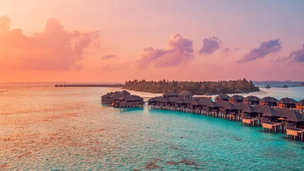 Vista aérea de bangalôs sobre águas cristalinas das Maldivas ao pôr do sol, com ilha tropical ao fundo.