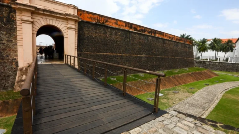 Ponte de madeira leva a uma passagem arqueada de pedra, parte de uma estrutura antiga. Pessoas à distância sugerem acesso par
